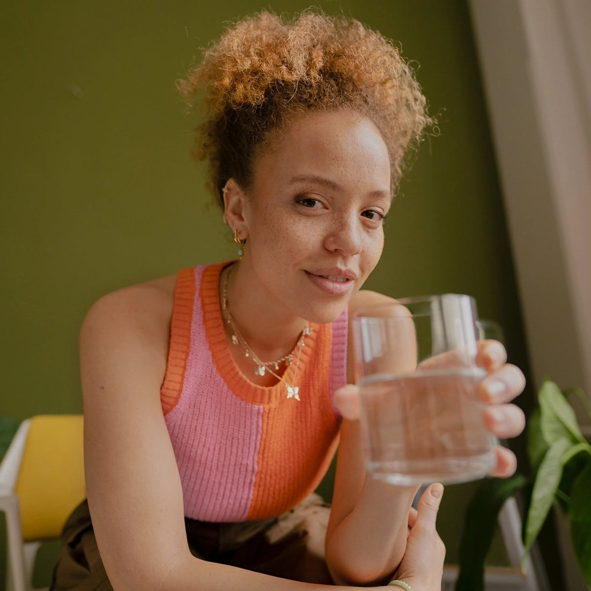 Woman in a pink top holding a glass of water in a home setting with plants.