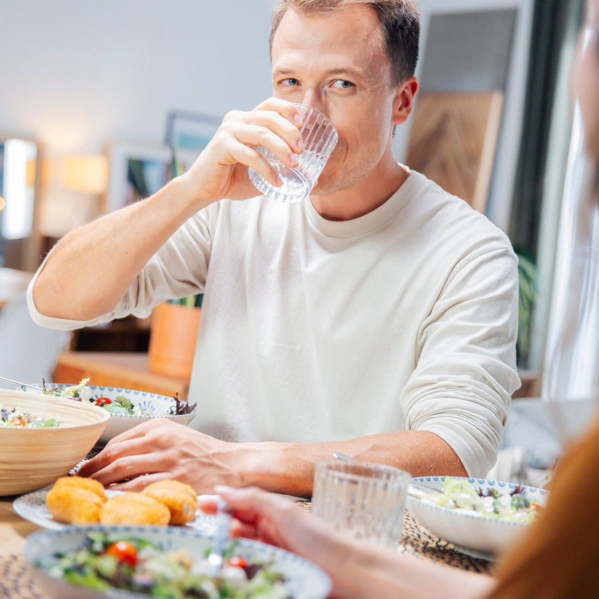 Un uomo che beve acqua a un tavolo da pranzo con del cibo e un'altra persona parzialmente visibile