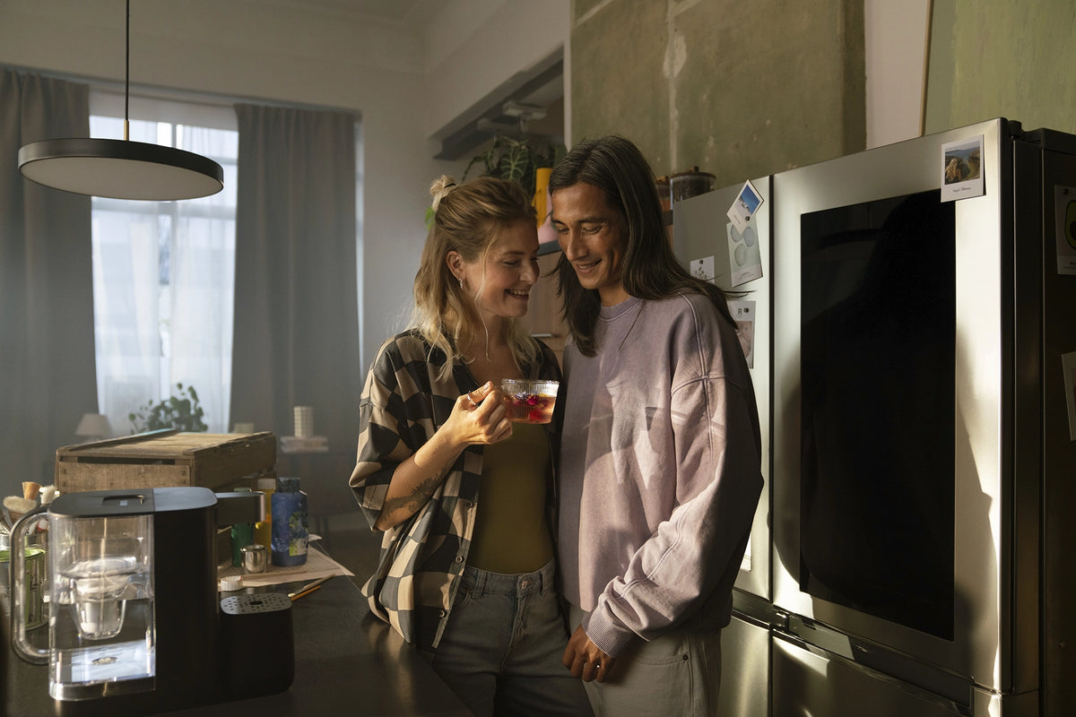 Two people standing in a kitchen next to a BRITA YOURCE mini water dispenser, with one person holding a glass cup of tea.